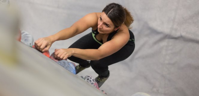 Eine Frau in Sportbekleidung klettert konzentriert eine Wand hinauf. Ihre beiden Hände sind nach oben gereckt und umfassen Kletterpunkte.