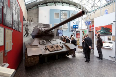 Im Museumsraum steht ein alter Panzer. Um ihn herum hängen viele Erklärtafeln. Erwachsene betrachten den Panzer. 