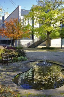 In einem Garten gibt es einen kleinen Teich mit Springbrunnen und Sitzmöbeln aus Holz. Hinter einigen Bäumen ist das Museum zu sehen.