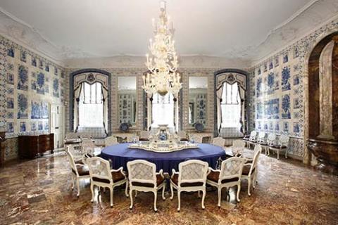 Der Sommerspeisesaal in Schloss Augustusburg. In der Mitte ein runder Tisch mit blauer Tischdecke mit vielen Stühlen. Von der Decke hängt ein prunkvoller Kronleuchter. Die Wand ist mit kunstvollem Porzellan vertäfelt.