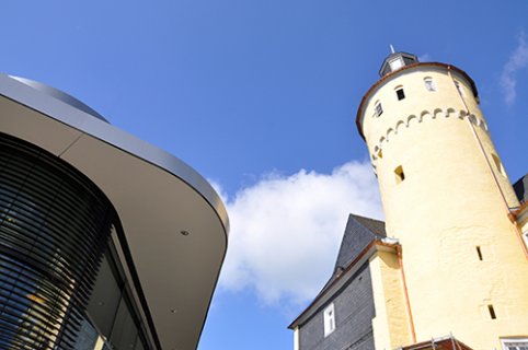 Ein gelber runder Turm des Schlosses ragt in den blauen Himmel. Neben dem Schloss sieht man Fenster und Dach eines zeitgenössischen Gebäudes.