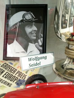 Auf einem Schwarzweiß-Foto ist ein Rennfahrer zu sehen. Darunter steht der Name Wolfgang Seidel. Rechts neben dem Foto steht ein glänzender Pokal. Links daneben liegt eine Urkunde auf der zu lesen ist: Großer Preis von Europa.