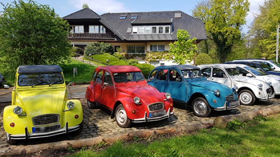 Vor einer großen Villa stehen ein gelbes, ein rotes, eins blaues und eine weißes Auto der Reihe Citroën 2CV - besser bekannt unter dem Namen "Ente" - geparkt.