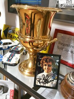 Auf einer Glasvitrine steht ein großer goldener Pokal. Vor ihm steht ein gerahmtes Foto von einem Rennfahrer mit Unterschrift. Drumherum parken einige weiße Modell-Rennautos.