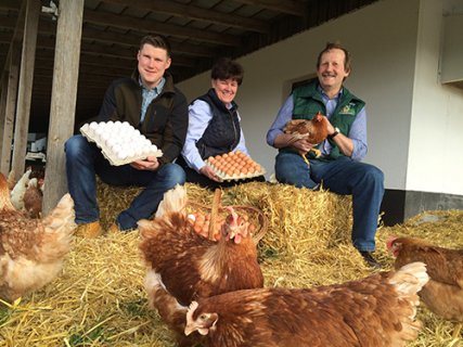 Drei erwachsene Menschen sitzen auf Strohballen vor einer Scheune. Sie sehen aus wie die Bäuerin, der Bauer und ihr erwachsener Sohn. Die Bäuerin und ihr Sohn präsentieren zwei große Paletten mit Eiern. Der Bauer hält eine Henne im Arm. Zu ihren Füßen picken weitere Hennen.