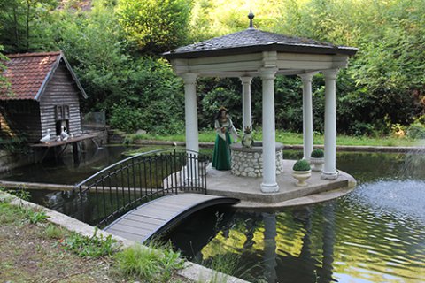 In einem Pavillon auf dem Wasser wird das Märchen vom Froschkönig dargestellt. Eine Prinzessinnenfigur in einem grün-weißen Kleid steht an einem Brunnen. Auf dem Brunnenrand sitzt ein Frosch mit einer Krone. Der Weiher ist umgeben von Bäumen und Wiesen. Seitlich ist ein Holzhaus zu sehen, vor dem eine Gänsefamilie sitzt.