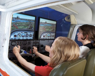 Zwei Kinder sitzen nebeneinander in einem Cockpit und simulieren auf ihren Displays einen Flug über Köln.