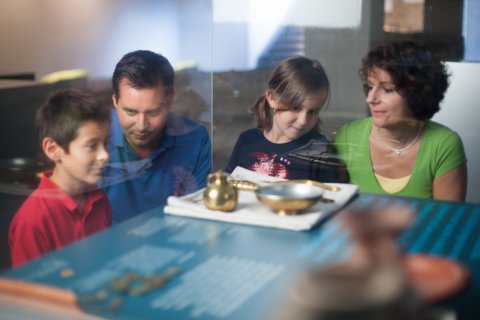 Eine Familie mit Mutter, Vater und zwei Kindern schauen in eine Vitrine. Dort sind unter anderem vergoldete Badeutensilien ausgestellt sowie ein kleines Kännchen und eine Schale.