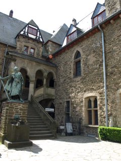 Eine Steintreppe führt hinauf zu einem von Rundbögen überdachten Eingang. Unten am Fuß der Treppe steht ein Ritterdenkmal aus Stein.