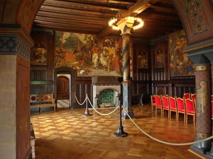 In einem Saal mit holzvertäfelten Wänden, Parkett und prächtigen Wandgemälden steht eine lange Tafel mit Stühlen aus rotem Samt. Neben einem großen Wandkamin führt ein Durchgang in einen anschließenden Raum. 