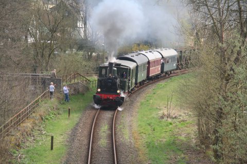 Eine Dampflok mit mehreren Waggons fährt durch eine Landschaft. Zwei Menschen stehen an den Gleisen und betrachten den durchfahrenden Zug.