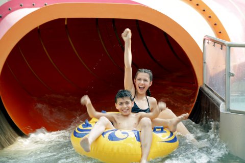 Aus der Öffnung einer orangenen Wasserrutsche kommen ein Mädchen und ein Junge auf einem gelben Schlauchboot gerutscht. Sie recken fröhlich die Arme nach oben.
