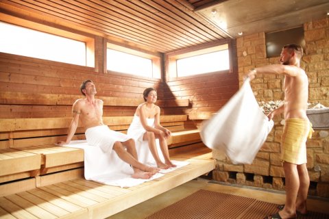 Eine Frau und ein Mann sitzen in einer großen, hellen Sauna. Ein zweiter Mann steht vor ihnen und schwenkt ein Handtuch beim Aufguss.