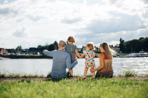 Blick von hinten auf eine junge Familie mit zwei Kindern. Sie sitzen auf einer Wiese direkt am Rhein. Der Mann und die Frau zeigen auf etwas, das auf dem Wsser oder am anderen Ufer sein muss. Die Kinder schauen in die gezeigte Richtung.