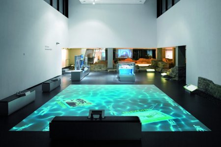 In einem modernen Museumsraum wird die Projektion eines türkis-leuchtenden Thermebeckens auf den Fußboden geworfen. Drumherum stehen verschiedene Exponate mit Erklärtafeln, wie zum Beispiel alte römische Mauerreste.