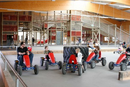 In einer Halle sitzen ein Mann und mehrere Kinder in roten Kett-Cars. Im Hintergrund ist eine Kletterlandschaft zu sehen.